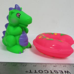Può includere: Una figura di drago giocattolo verde e viola con un occhio rosa e un dispositivo di galleggiamento rosa e verde. Il drago è seduto in posizione eretta e il dispositivo di galleggiamento è accanto. I giocattoli sono di plastica.