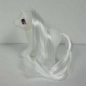 Könnte beinhalten: Ein weißes Einhorn-Pony-Spielzeug mit einer langen, fließenden weißen Mähne und einem weißen Schwanz. Das Pony hat ein violettes Auge und ein weißes Horn.