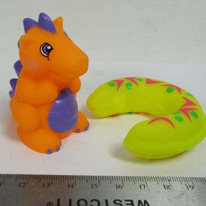 Può includere: Un giocattolo a forma di dinosauro arancione e viola con un cuscino per il collo giallo, verde e rosa a fantasia. Il dinosauro ha delle spine viola e una faccia amichevole. Il cuscino per il collo è curvo e decorato con motivi a zig-zag e a pois.