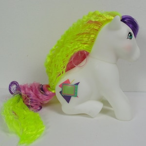 Puede incluir: Pony blanco con una crin y cola rizadas de color rosa y amarillo. El pony tiene un símbolo en su flanco de una pantalla de televisión con formas geométricas alrededor.