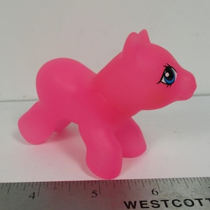 Puede incluir: Un pony de juguete de plástico rosa brillante con ojos azules. El pony está de pie sobre sus cuatro patas y tiene un acabado liso y brillante.