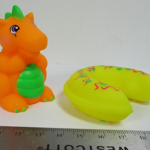 Può includere: Una figura di drago giocattolo arancione con un oggetto verde a forma di uovo. Accanto c'è un giocattolo giallo a forma di U con decorazioni rosa e verdi. I giocattoli sono su una superficie bianca.