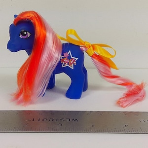 May include: A blue plastic pony toy with long orange and pink hair, featuring a white star symbol with roller skates on its flank and a yellow ribbon tied in its tail, positioned next to a metal ruler measuring over 11 inches.
