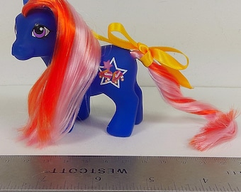 Könnte beinhalten: Ein blaues Kunststoff-Pony-Spielzeug mit langem orangefarbenem und rosafarbenem Haar, einem weißen Sternsymbol mit Rollschuhen auf der Flanke und einer gelben Schleife im Schweif, platziert neben einem Metalllineal von über 28 cm.