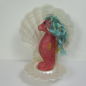 Op de afbeelding: Een roze en gele glitter zeepaardje speelgoed met blauw en iriserend haar, zittend in een witte schelp.