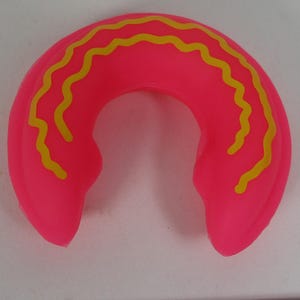 Puede incluir: Un anillo de goma rosa con líneas onduladas amarillas. El anillo tiene forma de herradura.