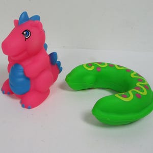 Può includere: Un giocattolo a forma di dinosauro rosa e blu e un giocattolo verde curvo con accenti gialli e rosa. Il dinosauro ha accenti blu e un occhio blu. Il giocattolo verde ha un design giallo ondulato con punti rosa.