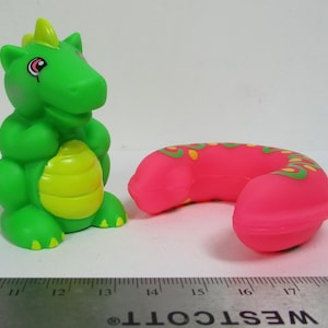 Può includere: Un giocattolo a forma di drago verde con dettagli gialli e un giocattolo rosa a forma di ferro di cavallo con disegni verdi e gialli. I giocattoli sono in materiale morbido, su una superficie bianca.
