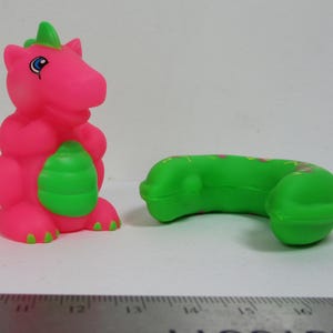 Può includere: Una figura di dinosauro giocattolo rosa e verde e una cornetta telefonica giocattolo verde. Il dinosauro ha una pancia verde e una cresta verde. La cornetta del telefono è verde con accenti rosa e gialli.