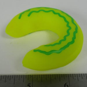 Puede incluir: Un anillo de goma amarillo brillante con líneas onduladas verdes. El anillo tiene forma de herradura.