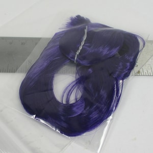 Könnte beinhalten: Ein Bündel tiefvioletter Kunsthaarverlängerungen, ordentlich in einer transparenten Plastiktüte verpackt. Das Haar hat einen leuchtenden Violettton mit leichtem Glanz. Ein silberner Draht sichert das Haar. Das Bild ist vor weißem Hintergrund.