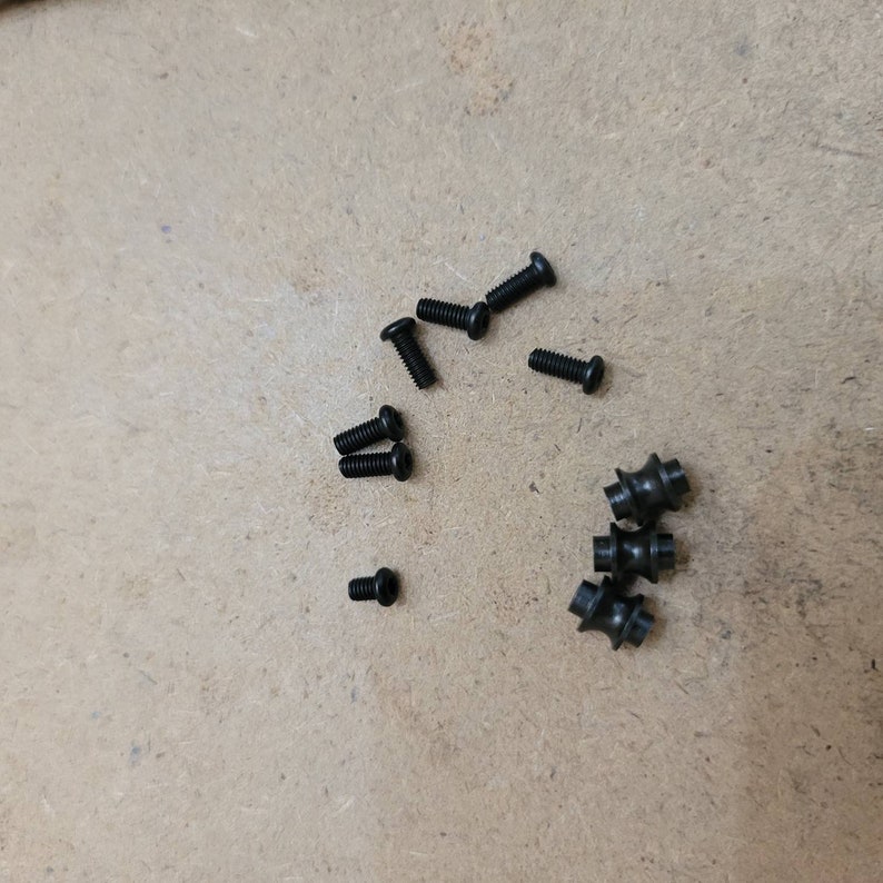 May include: A collection of small, black screws and fasteners scattered on a light brown surface. The screws have various head styles, including flat and rounded, and are likely used for assembly or repair.