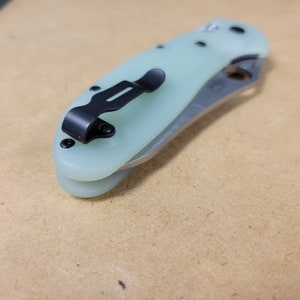May include: A light green folding knife with a black pocket clip and hardware. The blade is silver and partially visible. The knife is closed, showing the handle and clip. The handle has a smooth, rounded shape.