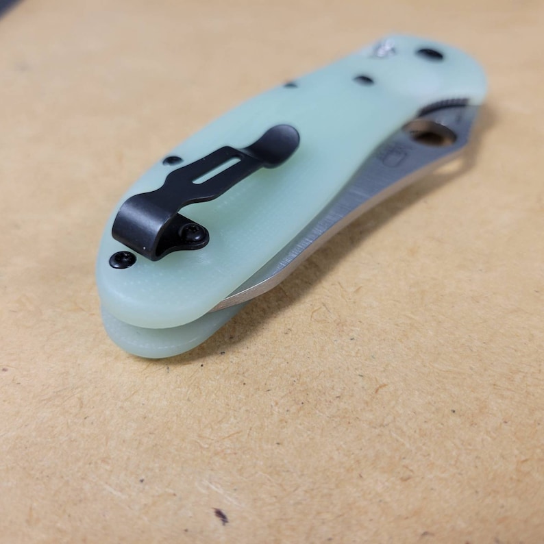 May include: A light green folding knife with a black pocket clip and hardware. The blade is silver and partially visible. The knife is closed, showing the handle and clip. The handle has a smooth, rounded shape.