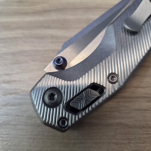 May include: Close-up of a silver folding knife with a partially visible blade. The handle has a textured, ridged design with a black button and several screws. A pocket clip is visible on the side.