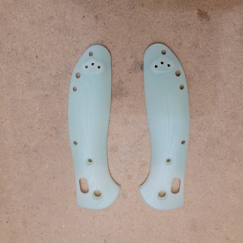 May include: Two pale green knife handle scales with multiple holes for attachment. The scales are curved and have a textured surface. The image is taken on a light brown surface.