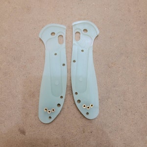 May include: Two translucent green knife handle scales with multiple holes and brass inserts. The scales are shaped to fit a folding knife, with a curved handle and a section for the blade. The scales are designed for a custom knife build.