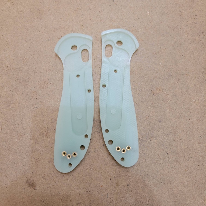 May include: Two translucent green knife handle scales with multiple holes and brass inserts. The scales are shaped to fit a folding knife, with a curved handle and a section for the blade. The scales are designed for a custom knife build.