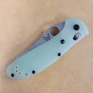 May include: A folding pocket knife with a light green handle and a silver blade. The knife has black hardware and the brand name "Benchmade" is visible on the blade. The knife is shown open, revealing the blade.