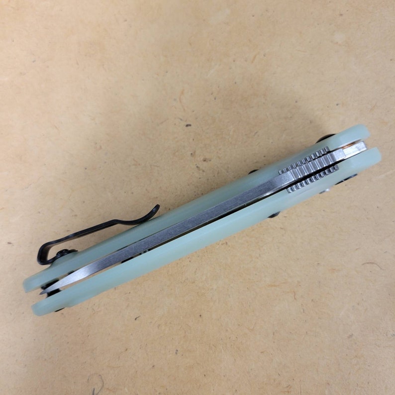 May include: A folding pocket knife with a light green handle and a black pocket clip. The blade is silver and the knife appears to be closed. The handle has a textured appearance.