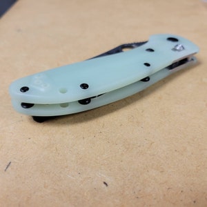 May include: A folding knife with a light green handle and a silver blade. The handle has black screws and a black clip. The knife is closed and resting on a light brown surface.