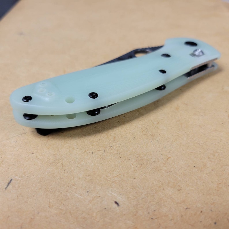 May include: A folding knife with a light green handle and a silver blade. The handle has black screws and a black clip. The knife is closed and resting on a light brown surface.