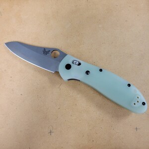 May include: A folding knife with a light green handle and a silver blade. The blade has a curved edge and a circular thumb hole. The handle has black accents and a pocket clip. The knife is designed for everyday carry.