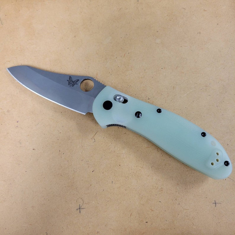 May include: A folding knife with a light green handle and a silver blade. The blade has a curved edge and a circular thumb hole. The handle has black accents and a pocket clip. The knife is designed for everyday carry.