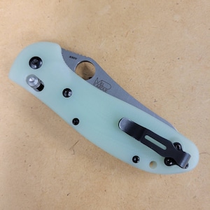 May include: A folding knife with a light green handle and a silver blade. The blade has the text "S30V" and "MIEL ARDUE". The knife also features a black pocket clip and black screws.