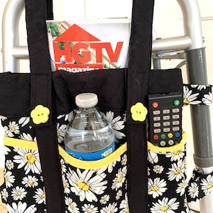 May include: Black and white daisy print tote bag with yellow trim and buttons. The bag is hanging on a walker and has a water bottle, remote control, and magazine inside.