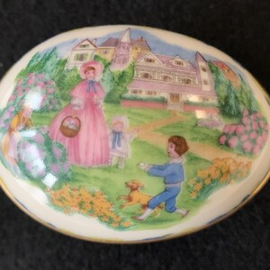Peut inclure: Une boîte à bijoux en porcelaine en forme d'œuf avec une scène peinte d'enfants jouant dans un jardin. La boîte représente une femme vêtue de rose, une jeune fille en robe bleue, un garçon en chemise et pantalon bleus et un chien jaune. L'arrière-plan comprend une grande maison avec un porche et une pelouse verdoyante.