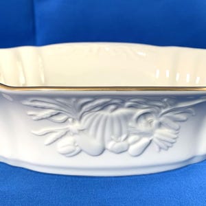 May include: A white oval serving bowl with a scalloped rim and gold trim. The bowl features a raised relief design of a floral and fruit arrangement on the front. The bowl is made of porcelain.