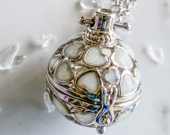 May include: A silver-toned locket necklace with a round, heart-patterned pendant. The pendant opens to reveal a small compartment. Clear, crystal-like stones and a small glass bottle are scattered around the necklace on a white surface.