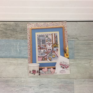 May include: A framed cross-stitch artwork titled "For the Bath" with a floral border. The artwork depicts a bathroom scene with a sink, mirror, and a rubber duck. A small ceramic container with a rose design and a sign that says "Be our Guest" are also present.