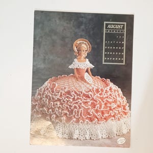 May include: A calendar page featuring a doll in a peach and white crocheted dress and hat. The doll is holding a fan. The calendar shows the month of August. The background is a dark gray color.