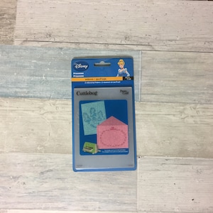May include: A Disney Princess Cuttlebug embossing folder set. The package features a blue and clear plastic case with a picture of Cinderella. The set includes two embossing folders and two embossing plates.