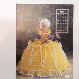 May include: A doll wearing a yellow crocheted dress with white lace trim and a matching bonnet. The doll holds a bouquet of purple and pink flowers. A calendar for May is in the upper right corner.