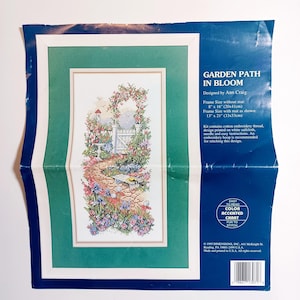 May include: A cross-stitch kit titled "Garden Path in Bloom" designed by Ann Craig. The image shows a framed cross-stitch design of a garden path with flowers, a white gate, and a birdhouse. The frame size is 13" x 21" (33x53cm).