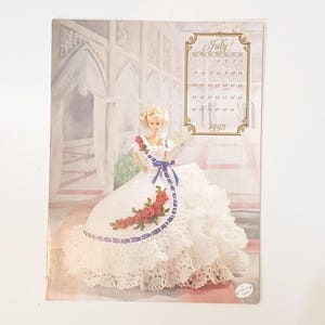 May include: A vintage print featuring a doll in a white lace gown with red rose accents and a blue ribbon. The print includes a July 1992 calendar. The doll has blonde hair and is posed in front of a soft, blurred background.