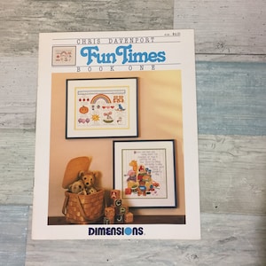 May include: A book cover for "Fun Times Book One" by Chris Davenport. The cover features two framed cross-stitch samplers with colorful designs. A basket with teddy bears and alphabet blocks are in the foreground.