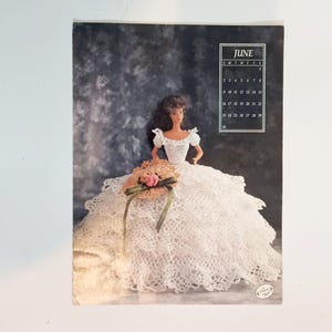 May include: A calendar page featuring a doll in a white crocheted dress and a matching hat with pink flowers. The calendar shows the month of June. The background is a dark, textured pattern.