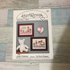May include: A cross-stitch pattern booklet titled "HeartStrings Yesterday's Child" featuring framed cross-stitch designs of a child's room, a heart-shaped ornament, and a white teddy bear. The booklet is from The Artists Collection.