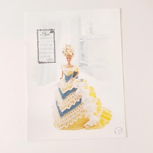 May include: A print featuring a Barbie doll in a yellow, white, and blue crocheted gown. The doll has blonde hair and is posed in front of a blurred background. The print includes a small calendar for April 1997.