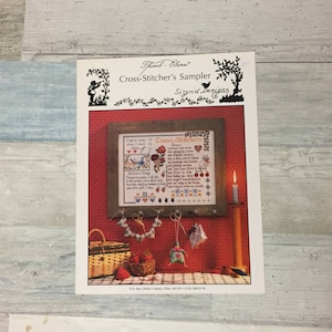 May include: A cross-stitch sampler pattern by Sigrid Designs, titled "Cross-Stitcher's Sampler." The image shows a framed cross-stitch piece with a poem, along with accessories like scissors and a basket. The pattern's title and designer's name are visible.