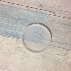 May include: A clear, round plastic lid with a small handle. The lid has a circular design in the center and is slightly translucent. The lid is likely for a container, and is sitting on a light-colored wooden surface.
