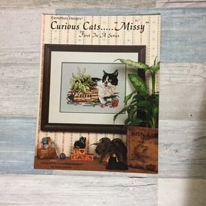 May include: A cross-stitch pattern titled "Curious Cats...Missy" by Canterbury Designs. The image features a framed cross-stitch of a black and white cat with a box of seeds. Additional elements include a small wooden box, a plant, and the text "I Love Cats."