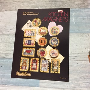 May include: A black leaflet featuring cross-stitch kitchen magnet designs. The magnets have gold-colored borders and depict various kitchen-themed images, including a watermelon, strawberry, and a pig. Text includes "KISS the COOK" and "HOME SWEET HOME".