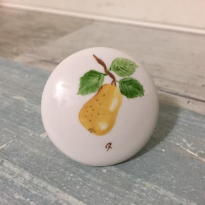 May include: White ceramic cabinet knob with a hand-painted yellow pear design. The pear has brown speckles, green leaves, and a brown stem. A small, stylized signature is visible near the bottom of the knob.