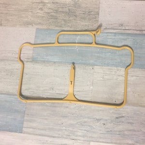 May include: A tan-colored plastic garment hanger with a unique design. The hanger has a rectangular frame with a handle at the top and a central support with the letter "T". The hanger is designed for a specific type of clothing.