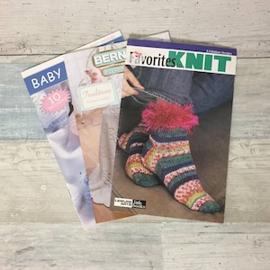 May include: A stack of knitting pattern books with titles including "BABY", "BERN", and "Favorites to Knit". The "Favorites to Knit" book features a colorful sock design. The books are on a light wood-grain surface.